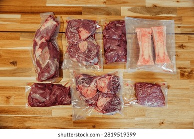 Top view of assorted raw beef cuts, organ meat, steaks, and marrow bones vacuum sealed in clear plastic bags on a wooden surface