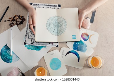 Top view of astrologer holding notebook with watercolor drawings and zodiac signs on cards on table