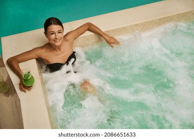 Top view of an attractive middle aged European woman in black swimsuit holding a freshly squeezed vitamin juice and looking at camera while getting relaxing hydromassage therapy in thermal pool at spa