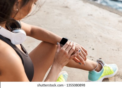 Top View Of An Attractive Young Fitness Woman Wearing Sportswear Sitting On A Fitness Mat Outdoors, Using Smartwatch