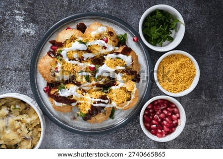 Top view of authentic Indian dahi papdi chaat topped with yogurt, tamarind chutney, sev, fresh coriander, and pomegranate seeds, served with ingredients on a rustic background. Popular traditional Indian street food snack, colorful vegetarian dish in flat lay style.