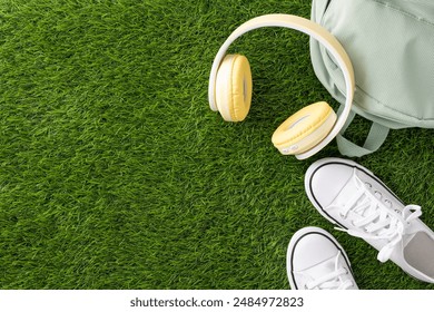 Top view of back to school essentials including yellow headphones, white sneakers, and a light green backpack placed on a green grass background