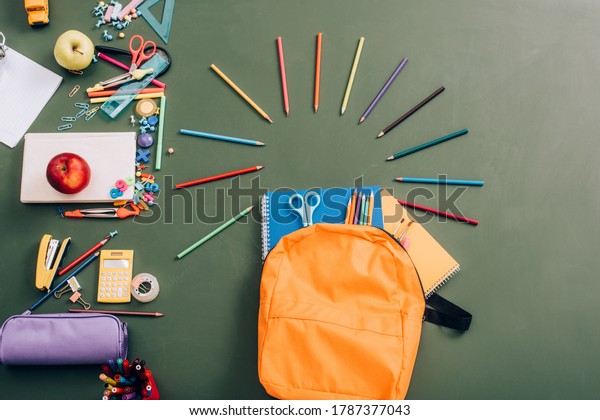 top view of backpack\
with notebooks, color pencils and scissors near school supplies on\
green chalkboard