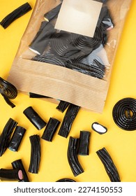 top view of a bag with black licorice jelly marmalade scattered over the background in the form of different tubes and sticks, on a yellow background.