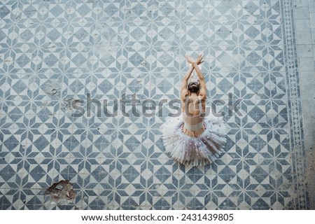 Top view of ballet dancer sitting in form bent forward and crossed arms against rustic blue and white tiled floor. Ballet diva performing in white leotard and tulle with pink flowers on a tiled floor.