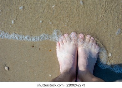 Top View Of Bare Men's Feet With Approaching Seawave On The Shore With Selective Focus, Low Angle Of Of A Man Standing On The Sand Beach, Activities And Recreation In Summer.