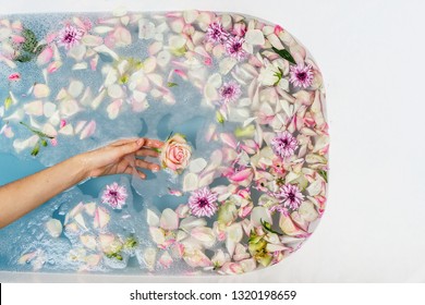 Top View Of Bath Filled With Blue Bubble Water, Flowers And Petals With Woman's Hand, Spa Or Selfcare Concept