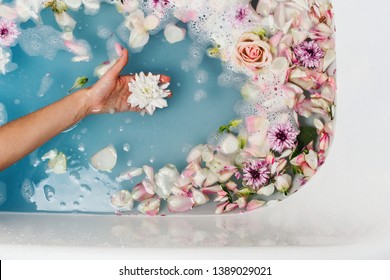 Top View Of Bath Filled With Blue Bubble Water And Petals With Woman's Hand Holding Flower, Spa Or Selfcare Concept