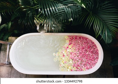 Top View Of Bath Tub Filling With Water With Flowers And Lemon Slices. Organic Spa Relaxation In Luxury Bali Outdoor Bathroom.