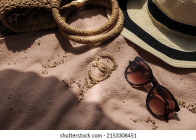 Top View Of Beach Accessories: Straw Hat, Summer Bag, Sunglasses, And Earrings On Bed Cover On Sand Beach On A Summer Day 