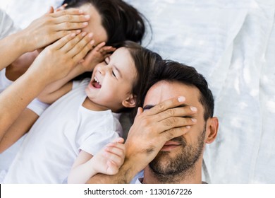 Top View Of Beautiful Family, Pretty Mother, Handsome Father And Cute Kid Playing Outdoors In Park. Horizontal Portrait Of Female And Male Closed Eyes With Hand Play Peek A Boo With Child. Parenting