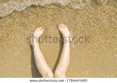 Top view of beautiful female feet with bright red pedicure on the sand of the beach.  The sea wave washes women's feet.  Relaxation and enjoyment during your seaside holiday.  Copy space.