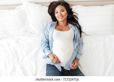 Top View Of A Beautiful Pregnant Asian Woman Lying On Bed At Home And Looking At Camera