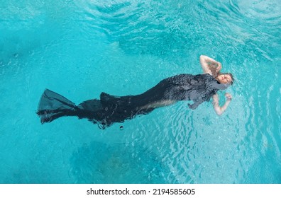 Top View To A Beautiful Sexy Young Woman Floating Like A Mermaid Fish, Wrapped In Black Cloth Dress In Turquoise Blue Water Of The Swimming Pool, Elegant Erotic Dream, Water Ballet, Copy Space