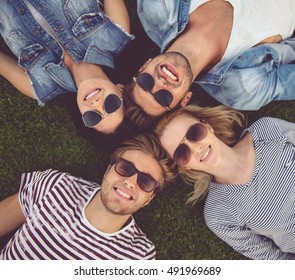 Top View Of Beautiful Stylish Friends In Sun Glasses Looking At Camera And Smiling While Lying On Grass