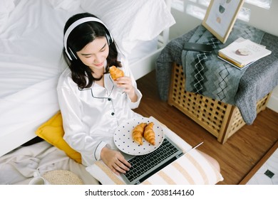 Top View Of Beautiful Young Asian Woman Sitting Next To The Bed Using Her Laptop And Enjoy Breakfast At Home. Small Business Owners Are Checking Orders Online. Concept For Freelancer Lifestyle.