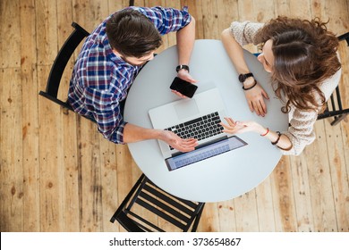 Top View Of Beautiful Young Couple Talking, Using Laptop And Holding Blank Screen Cell Phone At The Round Table In Cafe