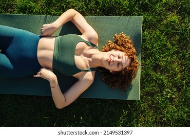 Top view of beautiful young happy sportive woman wearing green sports bra and blue yoga pant. Closed eyes and smiling while lying on yoga mat.