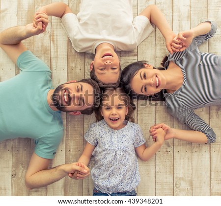 Top view of beautiful young parents, their cute little daughter and son holding hands, looking at camera and smiling, lying on wooden floor