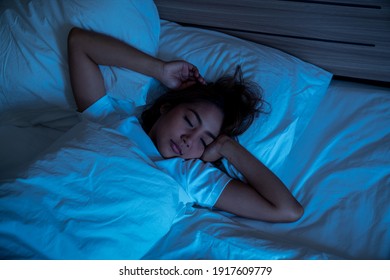 Top View Of Beautiful Young Woman Sleeping Cozily On A Bed In His Bedroom At Night. Blue Nightly Colors With Cold Weak Lamppost Light Shining Through The Window.