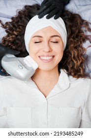 Top view of beautiful young woman with closed eyes wearing white headband while receiving facial treatment at wellness center. Cheerful female patient having face lifting procedure and smiling. - Shutterstock ID 2193485249