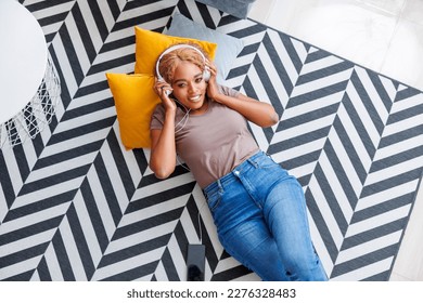 Top view of beautiful young woman lying on the living room floor wearing headset listening to the music and relaxing at home, enjoying her leisure time alone