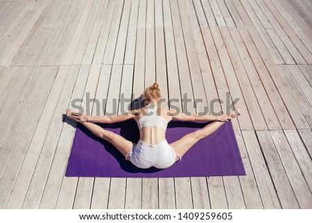 Top view. Beautiful young woman in white sportswear doing yoga outdoor on purple mat, wooden terrace. Girl sitting in Wide-Angle Forward Bend pose, Upavishtha Konasana. Yogi, sport and healthy concept