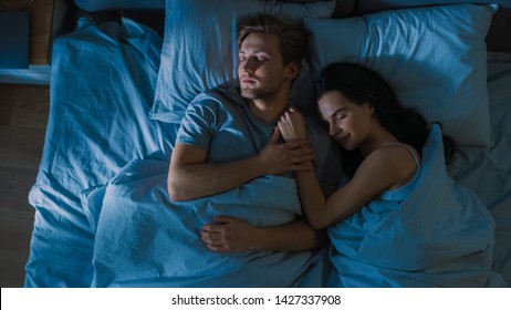 Top View Bed At Night: Attractive Young Couple Sleeping Together, Holding Each Other In Arms, Embracing. First Rays Of Morning Sun Illuminate Room Trough The Window