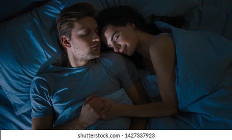 Top View Bed At Night: Attractive Young Couple Sleeping Together, Holding Each Other In Arms, Embracing. First Rays Of Morning Sun Illuminate Room Trough The Window