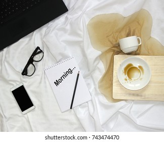 Top view of bed, notepad with black inscription morning, notebook, smart phone, glasses, spilled coffee from white cup on wood board. Home working mock up.
