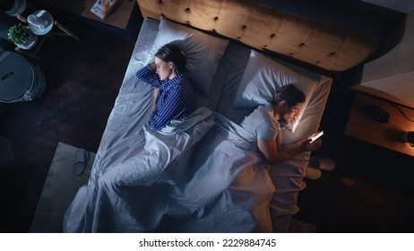 Apartamento de dormitorio con vista superior: Hombre usa Smartphone en cama por la noche cuando su pareja intenta dormir. Pareja Después De Pelear, Discusión. El mundo adictivo de los medios sociales, el desplazamiento de la condena, las noticias falsas.