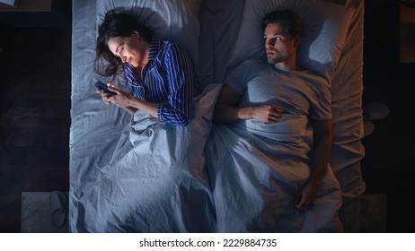 Top View Bedroom Apartment: Young Woman Uses Smartphone in Bed at Night When Her Male Partner Trying to Fall Asleep Beside. Couple Fight, Argue. Social Media, Doom Scrolling, Fake News Addiction
