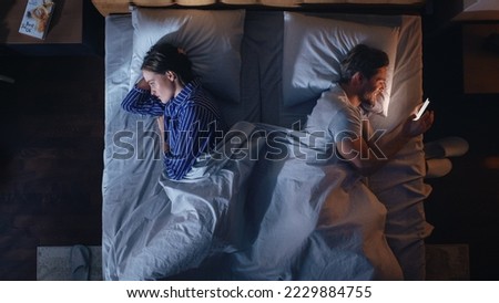 Top View Bedroom Apartment: Young Woman Uses Smartphone in Bed at Night When Her Male Partner Trying to Fall Asleep Beside. Couple Fight, Argue. Social Media, Doom Scrolling, Fake News Addiction