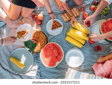 Top view of big family sitting on the picnic blanket in city park. They are eating boiled corn, apples, just cooced pie, peaches, pastries and watermelon. Family values and outdoor activities concept. - Powered by Shutterstock