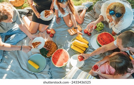 Top view of big family sitting on the picnic blanket in city park. They are eating boiled corn, apples, peaches, pastries and watermelon. Family values and outdoor activities concept - Powered by Shutterstock