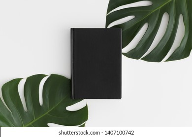 Top View Of A Black Book Mockup With Monstera Leaf Decoration On A White Table.