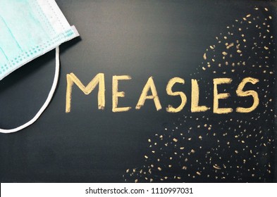 Top View Of A Black Chalkboard With The Medical Phrase Measles Written On It With Chalk And A Green Or Blue Medical Face Mask. Measles Outbreak, Vaccination Or Epidemic Concept