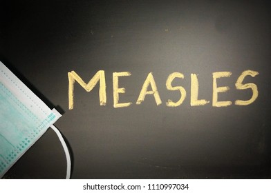 Top View Of A Black Chalkboard With The Medical Phrase Measles Written On It With Chalk And A Green Or Blue Medical Face Mask. Measles Outbreak, Vaccination Or Epidemic Concept