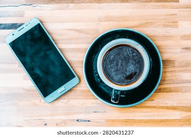 Top view of a black coffee cup and a smartphone on a wooden table text space.
Flat lay of hot black coffee espresso next to a smart phone on textured wooden surface.