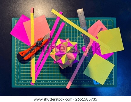 A top view of a black craft table with a cutting mat on it. Also, kusudama origami, colored papers, a metal ruler, and a utility knife create a cheerful and relaxed creative atmosphere.