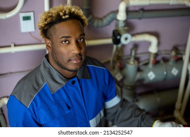 Top View Of A Black Worker In Overalls Standing In The Boiler Room And Holding A Suitcase With Special Tools. An African Man, A Professional In His Field, Looks At The Camera