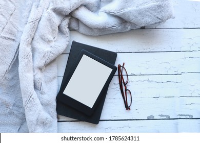 Top View Of A Blank Ebook, A Black Notebook On A White Rustic Wood Surface Next To A Fluffy Throw Blanket. Flataly Of An Ereader, Notebook And Blanket With Copy Space 