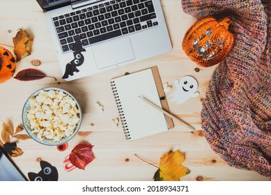 Top View Blank Notebook On Wooden Table With Laptop, Popcorn, Halloween Decor, Lighting Pumpkin, Autumn Leaves And Warm Plaid. Planning List Of Films For Halloween Movie Party. Flatlay. Copy Space