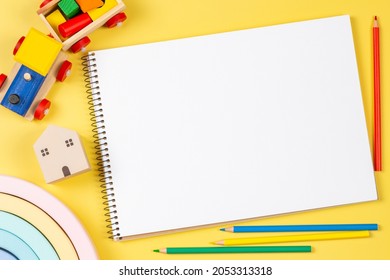 Top view to blank open sketchbook notebook with colored pencils and educational wooden kid toys and on yellow background. Early education, kindergarten, preschool, learn and play concept