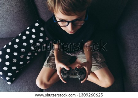 Top view of blond child with eyeglasses playing video games sitting on sofa  at home legs crossed Young student resting play on line with friends Domestic pastime challenge web Portrait from above