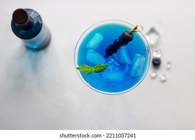 Top View Of Blue Cocktail In Martini Glass With Ice Cubes, Mint And Bluberries And Bottle Of Gin