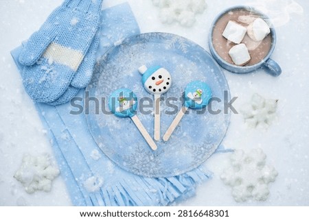 Top view of blue winter theme on snow with snowman cake pops, blue mittens, scarf, hot cocoa with marshmallows and snowflakes. Concept of winter, christmas, kids party and cozy.