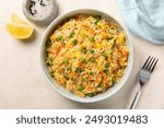 Top view of a bowl with fried rice with egg and vegetables, onion, bell pepper, carrot, green peas. Vegetarian food served in a light blue plate on a beige table.