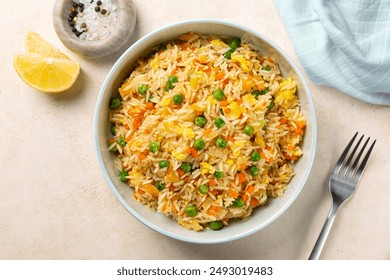 Top view of a bowl with fried rice with egg and vegetables, onion, bell pepper, carrot, green peas. Vegetarian food served in a light blue plate on a beige table.