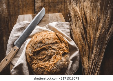 Top View Of A Bread Loaf And A Knife And A Cotton Rag On A Wooden Cutting Board, With Wheat Ears, On A Rustic Wooden Table